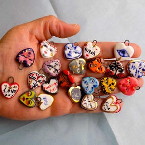 May include: A selection of heart-shaped pendants, each uniquely decorated with painted designs and text such as "Love" and "Witch." The pendants display a range of colours and patterns, including faces and eye motifs. Each has a small metal loop for attachment.