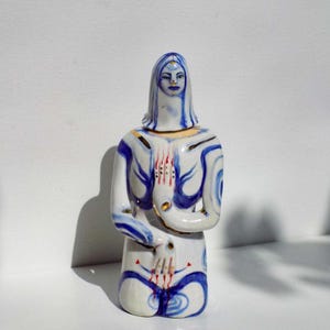 May include: A white ceramic figurine of a person kneeling with blue and gold painted details. The person has a blue face with a third eye painted on the forehead. The hands are painted with red and gold details.