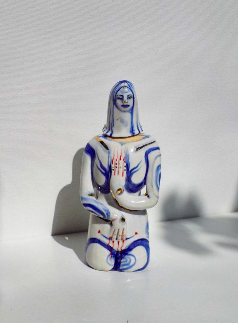 May include: A white ceramic figurine of a person kneeling with blue and gold painted details. The person has a blue face with a third eye painted on the forehead. The hands are painted with red and gold details.