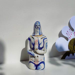 May include: A white ceramic figurine of a woman with blue and gold painted details. The woman is kneeling with her hands clasped in front of her.