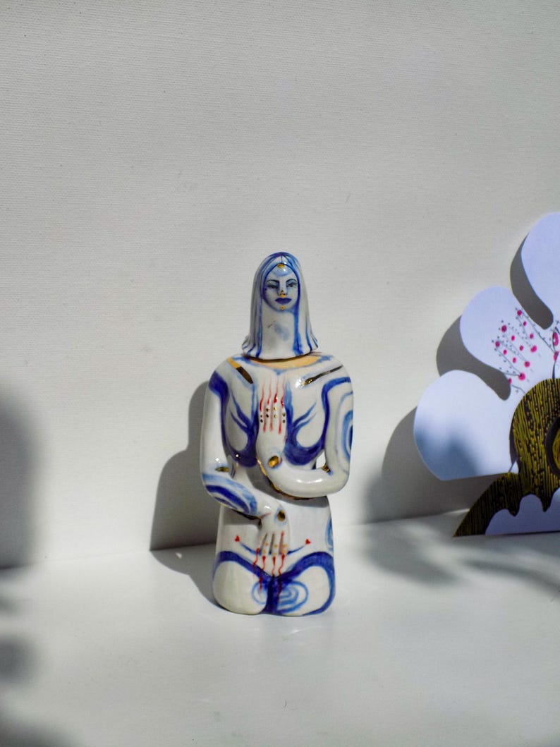 May include: A white ceramic figurine of a woman with blue and gold painted details. The woman is kneeling with her hands clasped in front of her.