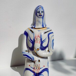 May include: A ceramic figurine of a woman kneeling with her hands clasped in prayer. The figurine is painted in shades of blue, white, and gold, with red accents. The woman's face is painted with a serene expression.