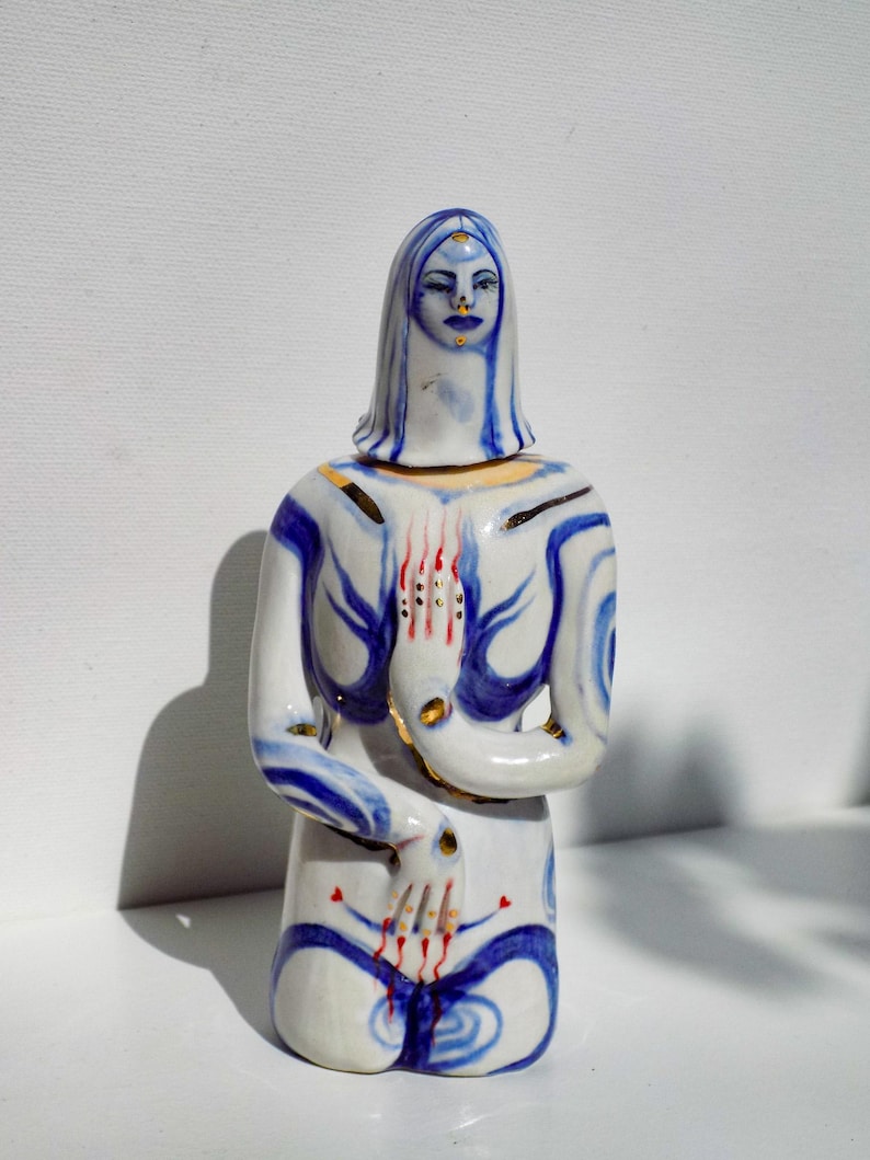 May include: A ceramic figurine of a woman kneeling with her hands clasped in prayer. The figurine is painted in shades of blue, white, and gold, with red accents. The woman's face is painted with a serene expression.