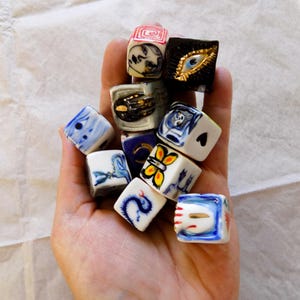 May include: A selection of small, square ceramic dice, each hand-painted with unique designs. The dice feature various colours, including blue, red, gold, and black, with images of eyes, faces, and abstract patterns. The dice are held in a hand.