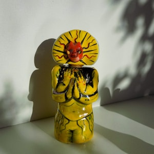 May include: A yellow ceramic figurine with a red face and a yellow sun-like crown. The figurine is wearing a yellow and black patterned outfit and has its hands clasped in front of it.