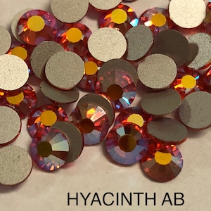 May include: A close-up of a pile of small, round, iridescent red rhinestones with a gold backing. The rhinestones are scattered on a white surface. The text "HYACINTH AB" is visible at the bottom of the image.