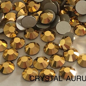 May include: A close-up image of gold-coloured rhinestones. The rhinestones are scattered on a white surface. The text "CRYSTAL AURUM" is printed in black at the bottom of the image.