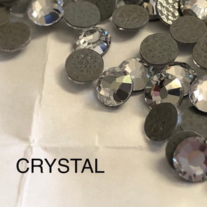 Puede incluir: Primer plano de un grupo de piedras de cristal transparentes con engastes plateados. Las piedras están esparcidas sobre una superficie blanca. La palabra "CRYSTAL" está impresa en negro debajo de las piedras.