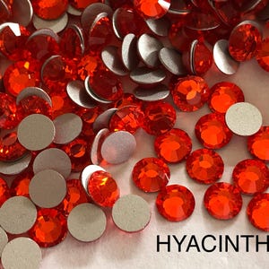Puede incluir: Una imagen de primer plano de una gran cantidad de pequeños diamantes de imitación redondos de color naranja con respaldo adhesivo. Los diamantes de imitación están esparcidos sobre una superficie blanca. La palabra "HYACINTH" está impresa en letras negras en la parte inferior de la imagen.
