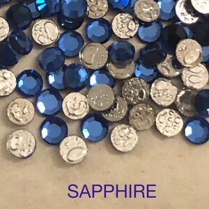 May include: A close-up of a pile of sapphire blue rhinestones with silver settings. The rhinestones are scattered on a light brown surface. The word "SAPPHIRE" is printed in purple below the rhinestones.