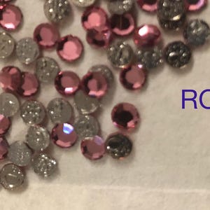 May include: A close-up of a variety of pink and silver rhinestones. The rhinestones are scattered on a white surface. The word "ROSE" is written in blue letters on the right side of the image.