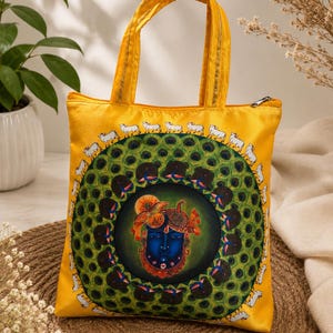 May include: A bright yellow tote bag featuring a central circular design with a blue face illustration surrounded by a green peacock feather pattern. A border of small white cows encircles the central motif. The bag has two yellow handles.