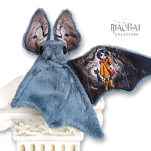 Può includere: Un giocattolo a forma di pipistrello di peluche blu, con grandi orecchie con un disegno di venature scure. Un'ala mostra un'illustrazione colorata di una ragazza con un cappotto giallo e un gatto nero. Il giocattolo è esposto su una colonna decorativa bianca. Il testo "MAOBAT CREATIONS" è visibile.