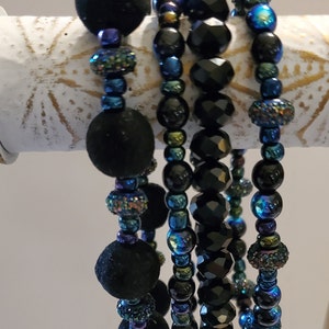May include: Five black beaded bracelets with iridescent blue and green accents. The bracelets are stacked on a white surface.