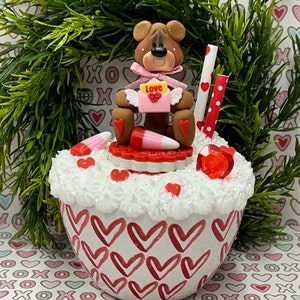 May include: A white ceramic mug with a red heart pattern, filled with white frosting and red and pink candies. A brown teddy bear with pink wings holds a pink envelope with the word "Love" on it. The mug is sitting on a background of pink, red, and white hearts.