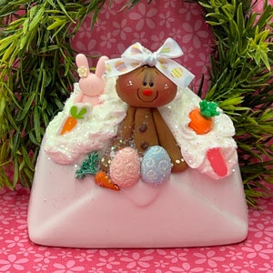 May include: A white envelope with a gingerbread person holding Easter eggs and a bunny rabbit. The envelope is decorated with white frosting and glitter.
