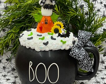 May include: Black ceramic cauldron mug with a white "BOO" inscription. The mug is decorated with a white ghost, a pumpkin, and a sunflower. The rim of the mug is covered in white faux snow and black bats.