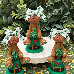 May include: Three brown gingerbread people wearing green hats and white bows with shamrock designs. Each gingerbread person is holding a green shamrock.