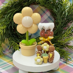 May include: A decorative display featuring a large yellow flower in a pot, a gingerbread man with a white hat and yellow bow, and several small flower and ice cream cone figurines. The display sits on a white, round, elevated platform.