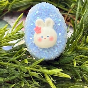 May include: A blue glittery Easter egg with a white bunny face and a pink flower. The bunny has a happy expression and is surrounded by green foliage.