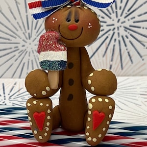 May include: A gingerbread person figurine wearing a red, white, and blue bow and holding a red, white, and blue popsicle. The figurine is sitting on red and blue striped straws.