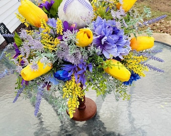 Peut inclure: Composition florale décorative avec une figurine d'oie blanche portant un chapeau de paille et un nœud vichy bleu. L'arrangement comprend des tulipes jaunes, de la lavande violette et des fleurs bleues et jaunes, sur un support en bois.