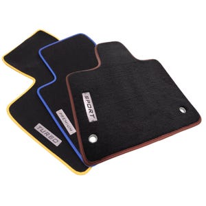 May include: Three black car floor mats with distinct colored edges in yellow, blue, and brown. Each mat features a rectangular metal badge with the text TURBO, PREMIUM, or SPORT, and includes two circular metal grommets for secure installation.