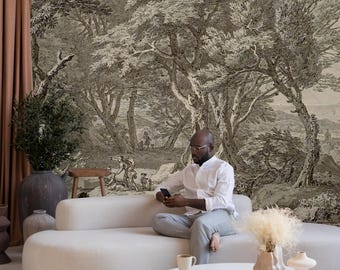 May include: A man sits on a light gray couch in front of a wall mural depicting a black and white forest scene. The couch is a modern design with a rounded shape. A white coffee table with four legs sits in front of the couch. The table has a book and a cup on it. There are two decorative vases on the table, one with a tall, thin, white shape and the other with a shorter, wider, beige shape.