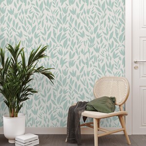 Puede incluir: Una habitación con papel pintado con un patrón de hojas verde claro sobre un fondo blanco. Una silla de madera con asiento y respaldo de mimbre tiene un cojín verde y una manta gris oscuro. Hay una planta en maceta y libros.