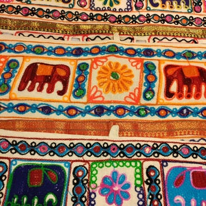 May include: A white fabric strip with colorful embroidered designs. The designs include elephants, flowers, and geometric patterns. The fabric is decorated with a gold trim.