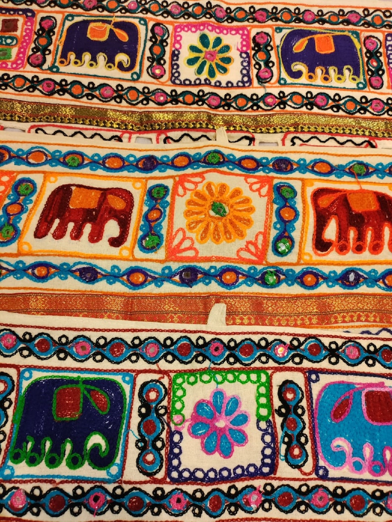 May include: A white fabric strip with colorful embroidered designs. The designs include elephants, flowers, and geometric patterns. The fabric is decorated with a gold trim.