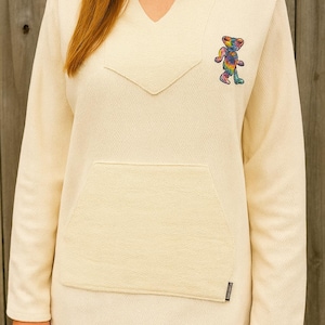 May include: Cream-colored hoodie with a V-neck and a kangaroo pocket. The long-sleeved top features a colorful tie-dye bear graphic on the upper left chest. The fabric has a subtle herringbone pattern.