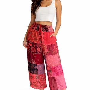 May include: A woman wearing a white tank top and patchwork palazzo pants. The pants feature a variety of red, pink, and purple fabrics with floral and geometric patterns. The pants have pockets and are cropped at the ankle.