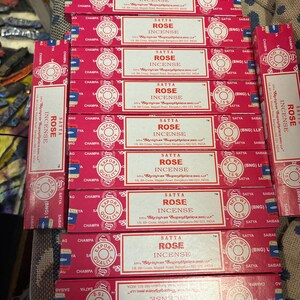 May include: A stack of ten pink and white boxes of Satya Rose Incense. Each box has a white label with the text "Satya Rose Incense" and the company's name and address. The boxes are stacked on a brown surface.