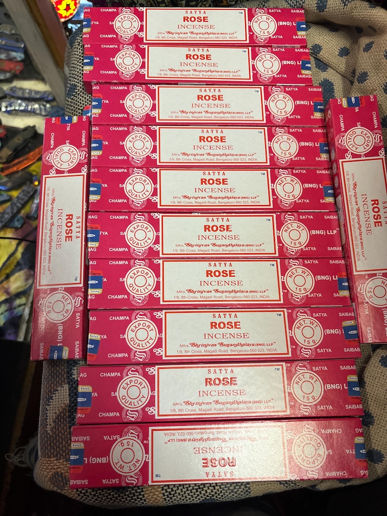 May include: A stack of ten pink and white boxes of Satya Rose Incense. Each box has a white label with the text "Satya Rose Incense" and the company's name and address. The boxes are stacked on a brown surface.