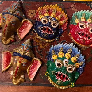 May include: Five colorful decorative masks, including a gold elephant head mask, a blue and red demon mask, a red and green demon mask, a green and yellow demon mask, and a blue and yellow demon mask.