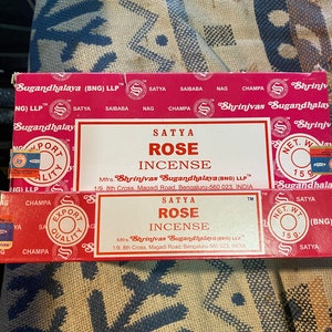 May include: A red and white box of Satya Rose incense. The box has a gold border and the text "Satya Rose Incense" is printed in black. The box also includes the manufacturer's name and address: "Mfrs. Shrinivas Sugandhalaya (BNG) LLP 1/9, 8th Cross, Magadi Road, Bengaluru-560 023, INDIA".