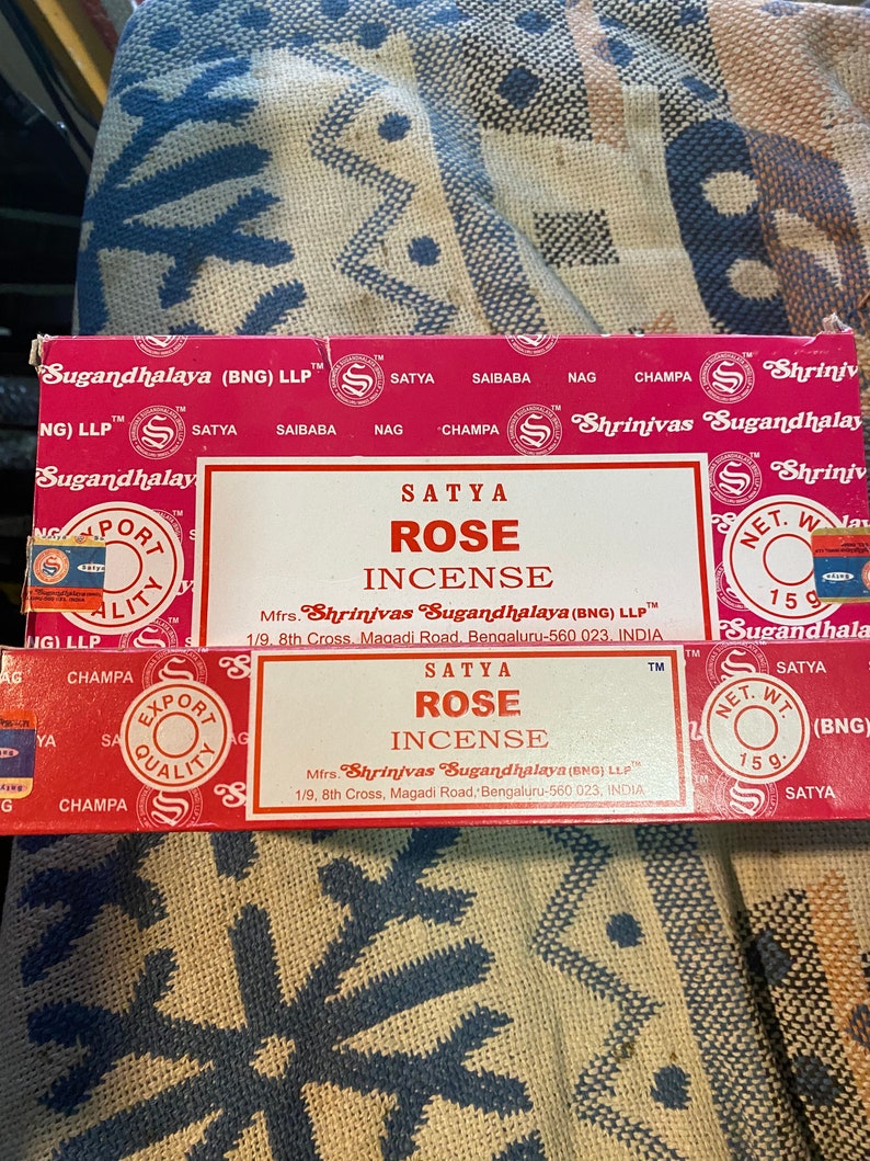 May include: A red and white box of Satya Rose incense. The box has a gold border and the text "Satya Rose Incense" is printed in black. The box also includes the manufacturer's name and address: "Mfrs. Shrinivas Sugandhalaya (BNG) LLP 1/9, 8th Cross, Magadi Road, Bengaluru-560 023, INDIA".