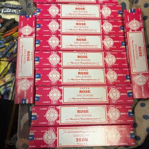 May include: A stack of red and white boxes of Satya Rose Incense. The boxes are labeled with the brand name, product name, and weight. The boxes are stacked on a brown surface.