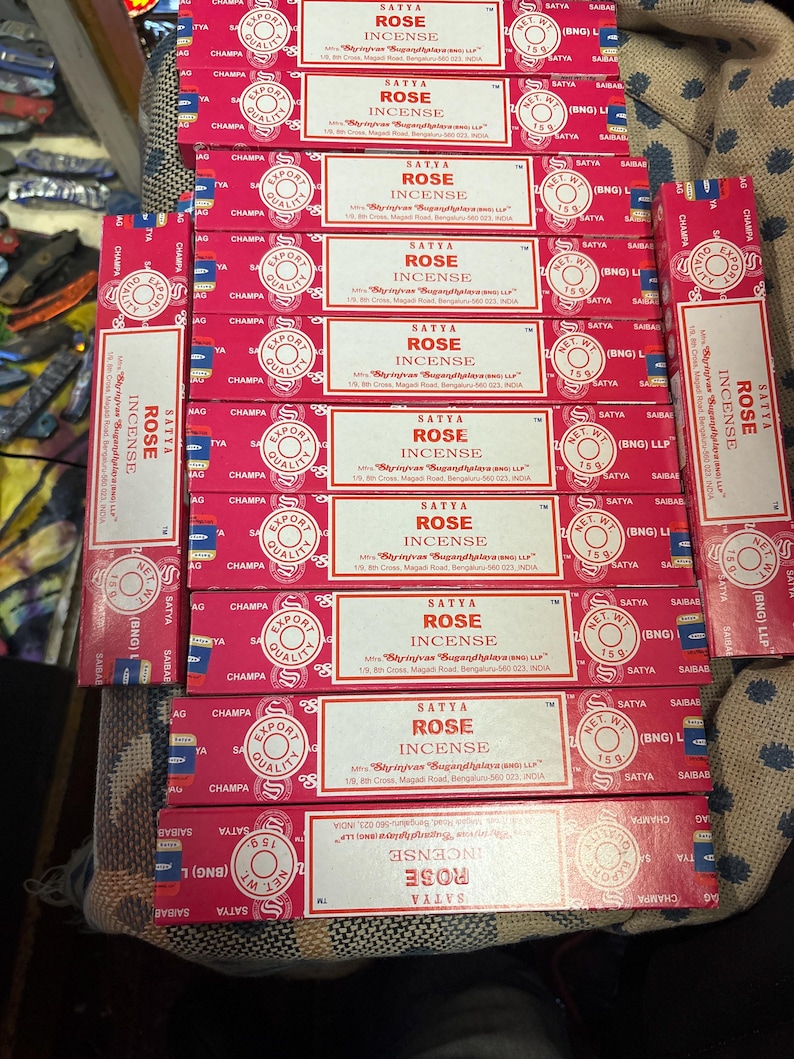 May include: A stack of red and white boxes of Satya Rose Incense. The boxes are labeled with the brand name, product name, and weight. The boxes are stacked on a brown surface.