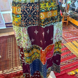 May include: A long, colorful patchwork skirt with a drawstring waist. The skirt is made of different fabrics with various patterns, including floral, geometric, and paisley designs. The skirt is a vibrant mix of red, orange, yellow, green, blue, and purple.