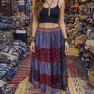 May include: A woman wearing a black crop top and a tiered maxi skirt in shades of purple and burgundy. The skirt has a floral pattern. She is wearing sandals and jewelry. The background shows a shop with textiles and rugs.
