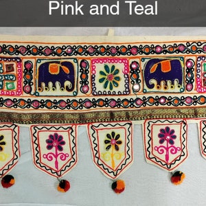 May include: A white cotton door hanging with a pink and teal embroidered design. The design features elephants, flowers, and geometric patterns. The hanging has a fringe border with tassels.