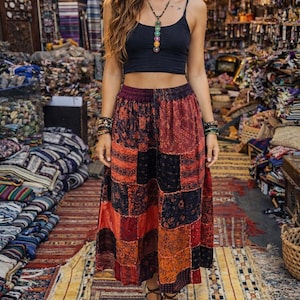 May include: A woman wearing a black crop top and a patchwork skirt in shades of red, orange, and black. She is wearing sandals and jewelry. The background shows a shop with textiles and hanging lanterns.