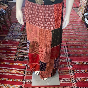 Puede incluir: Una falda larga de patchwork en tonos rojos, rosas y marrones, exhibida en un maniquí. La falda presenta una variedad de paneles de tela estampada, con una cintura negra y un cordón. La falda es larga y tiene un estilo bohemio.
