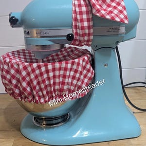 May include: A light blue KitchenAid Artisan stand mixer with a red and white checkered bow and bowl cover. The mixer is on a wooden surface. The text "Mini Homesteader" is visible on the mixer.