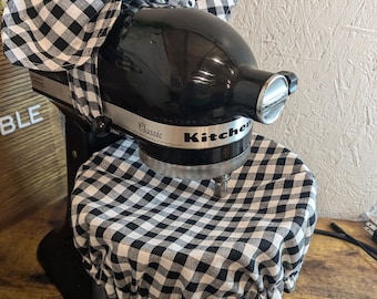 May include: Black and white checkered fabric cover for a KitchenAid stand mixer. The cover has a bow on top and the mixer has the word "Classic" printed on it.