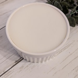 May include: A white ceramic ramekin with a fluted exterior and a smooth, round interior. The ramekin is filled with a white substance, possibly a dessert or dip. The background is a light-coloured wooden surface.