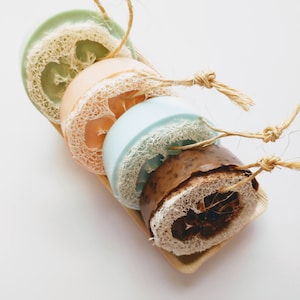 May include: Four round bath poufs with natural fibers, tied together with twine. The poufs are in shades of green, orange, blue, and brown.