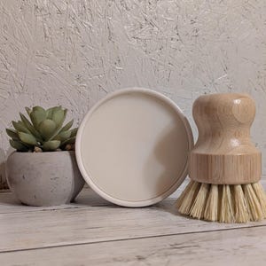 May include: A small succulent plant in a grey concrete pot, a white round soap dish, and a wooden dish brush with natural bristles are arranged on a light wood surface. The background is a textured white wall.